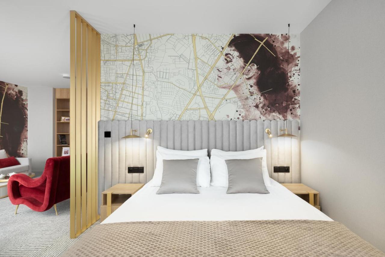 Royal Tulip Warsaw Centre Varsóvia Exterior foto The photo shows a stylish hotel room with a modern aesthetic. It features a bed with a light-colored duvet and several decorative pillows in shades of gray. Behind the bed is a unique headboard with vertical striped design. Above the headboard, there