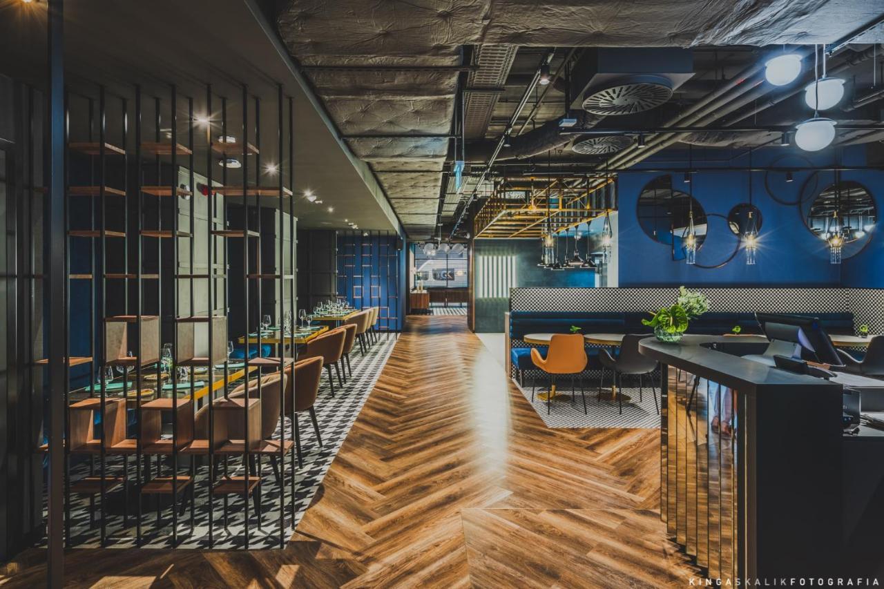 Royal Tulip Warsaw Centre Varsóvia Exterior foto The image shows a modern, stylish restaurant interior. There is a long wooden table with chairs on one side, and a bar area with a shiny counter on the other. The flooring features a herringbone pattern, adding warmth to the space. The walls are pain
