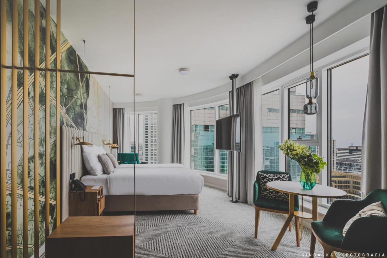 Royal Tulip Warsaw Centre Varsóvia Exterior foto The photo shows a modern hotel room with large windows that let in natural light. The decor features a stylish and contemporary design, including a bed with a white comforter and elegant headboard. There's a wooden nightstand beside the bed and a sma