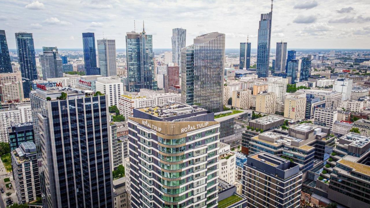 Royal Tulip Warsaw Centre Varsóvia Exterior foto The photo shows a panoramic view of a modern city skyline. In the foreground, there are several tall buildings with contemporary architectural designs. One building prominently features the name "Royal Tulip." The background is filled with numerous s
