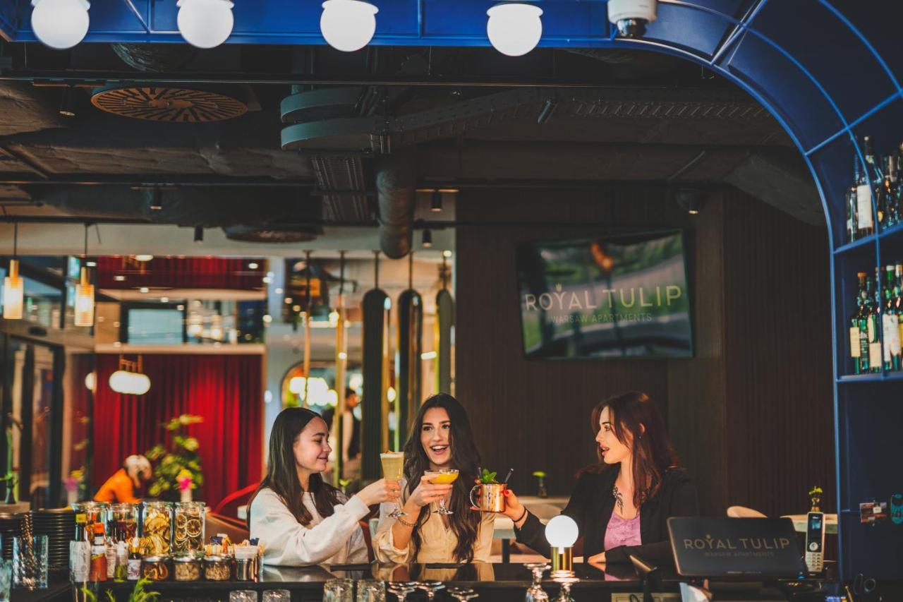 Royal Tulip Warsaw Centre Varsóvia Exterior foto The photo depicts three women gathered around a bar, enjoying drinks. They are smiling and engaged in conversation, exuding a festive and cheerful atmosphere. The bar has a modern design, with colorful decor and lighting. In the background, there is 