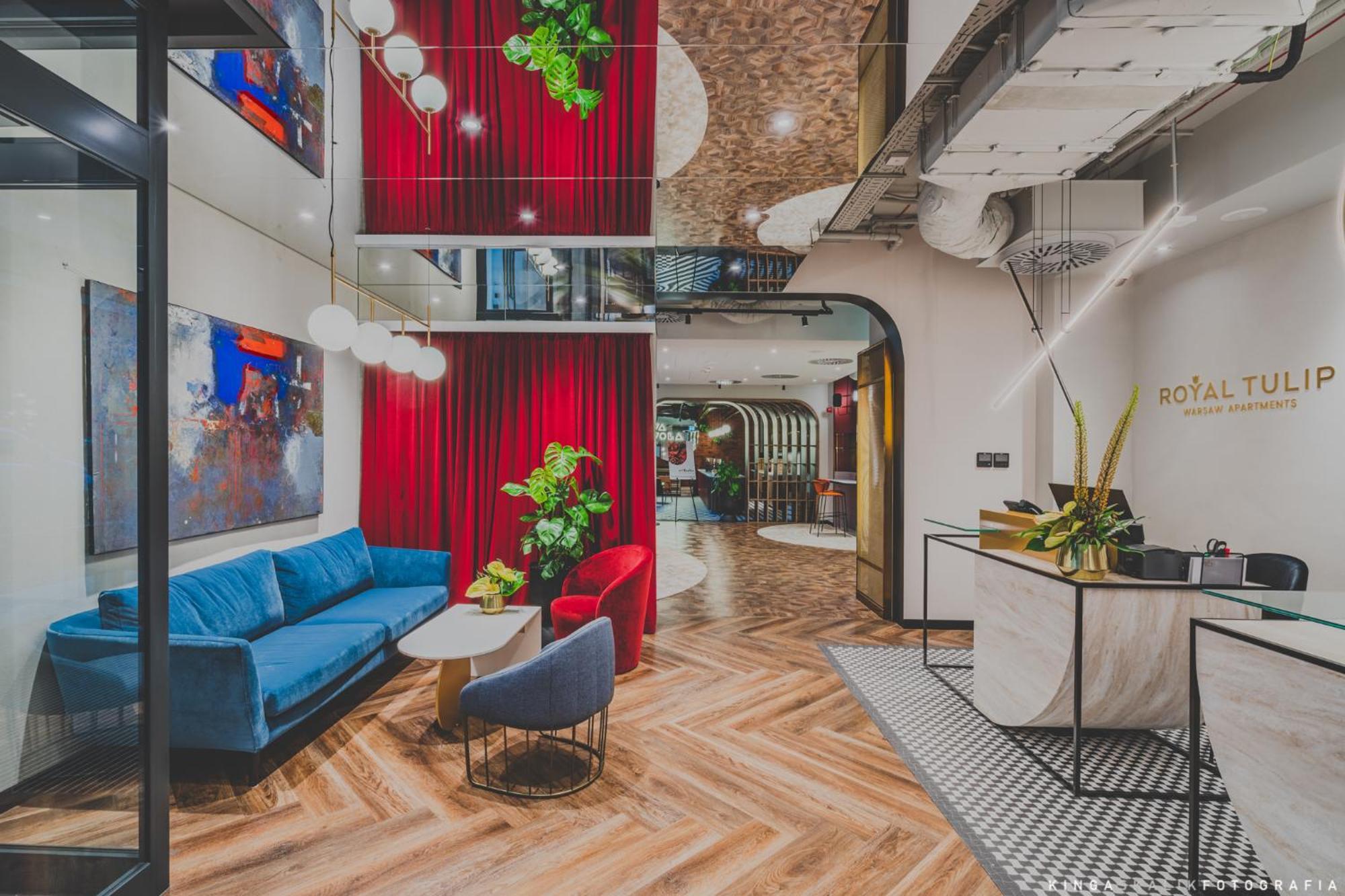 Royal Tulip Warsaw Centre Varsóvia Exterior foto The photo showcases a modern interior space, likely a lobby or lounge area. It features a mix of textures and colors, including a blue couch and red accent chairs. Behind the seating area, there are vibrant red curtains and greenery that adds a touch
