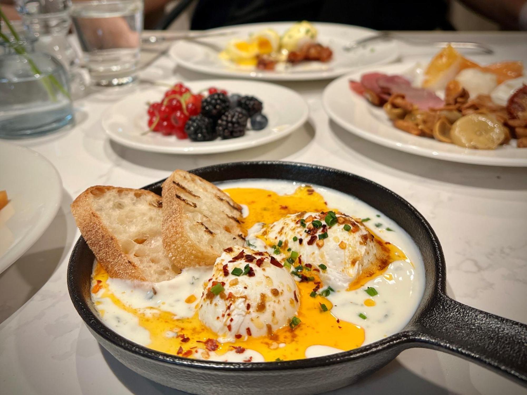 Royal Tulip Warsaw Centre Varsóvia Exterior foto The photo showcases a delicious breakfast spread. In the foreground, there is a cast-iron skillet containing three poached eggs nestled in a creamy sauce, garnished with herbs and red pepper flakes. Next to the skillet, there are slices of toasted br