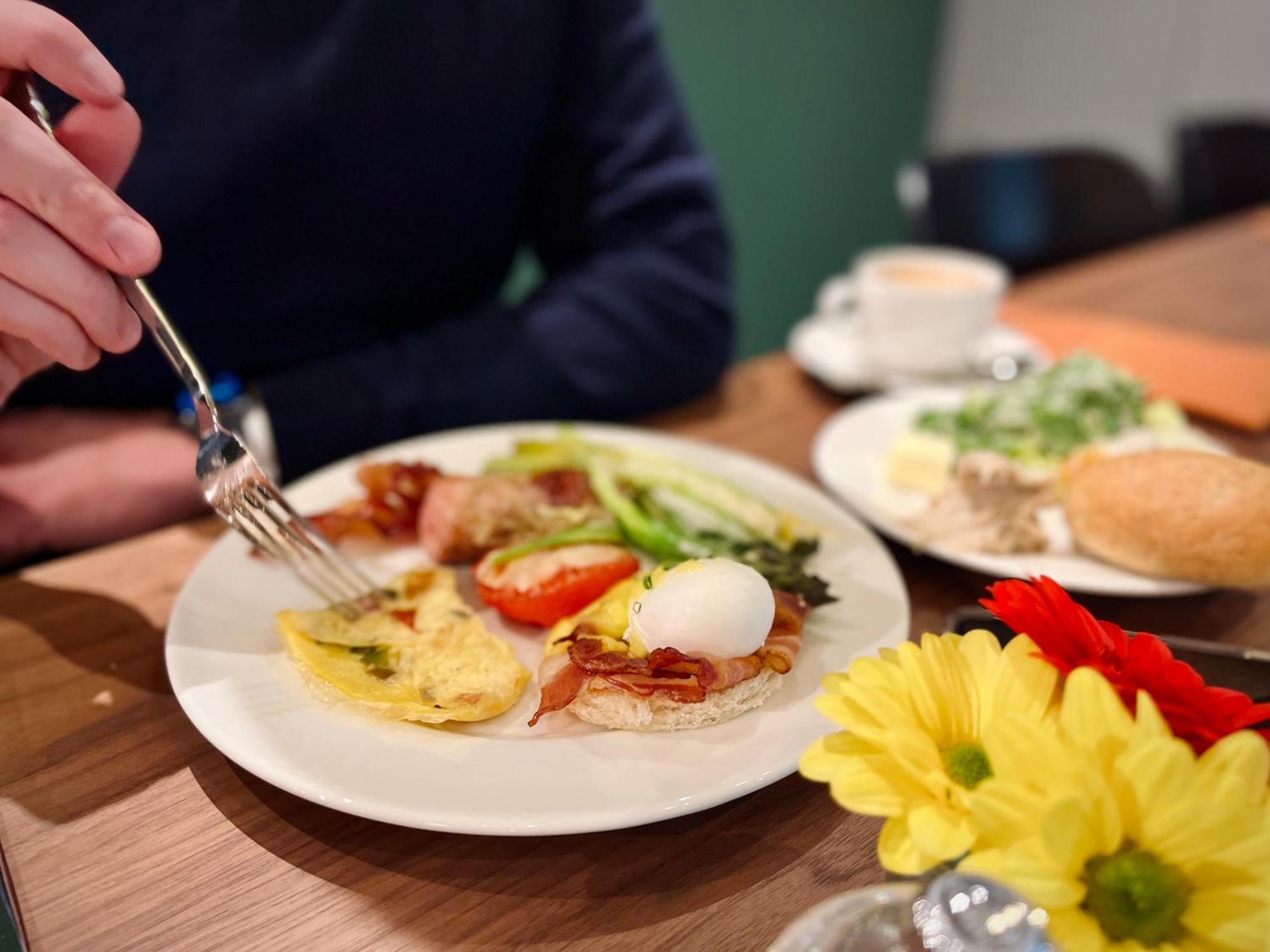 Royal Tulip Warsaw Centre Varsóvia Exterior foto The photo displays a breakfast plate with various items. There's an omelet, a poached egg, bacon, sliced tomatoes, and some greens. A hand is reaching for the food with a fork. Additionally, there are flowers on the table and a small cup of coffee. T