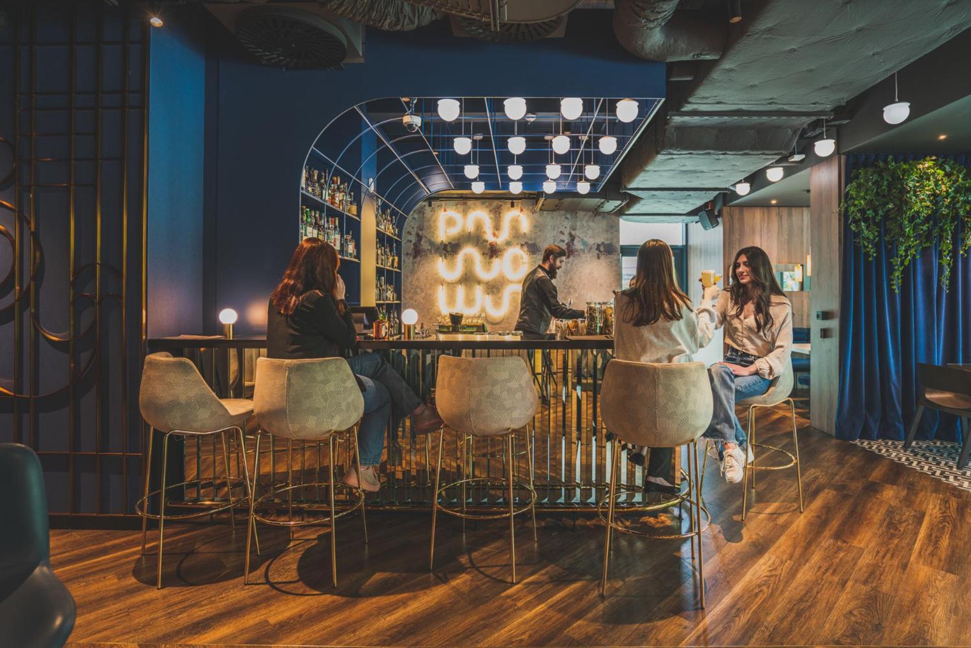 Royal Tulip Warsaw Centre Varsóvia Exterior foto The photo depicts a modern, stylish lounge or bar interior. There are several people sitting at a curved bar with high-back stools, engaged in conversation. The bar area is well-lit with decorative lighting that creates a warm atmosphere. In the back