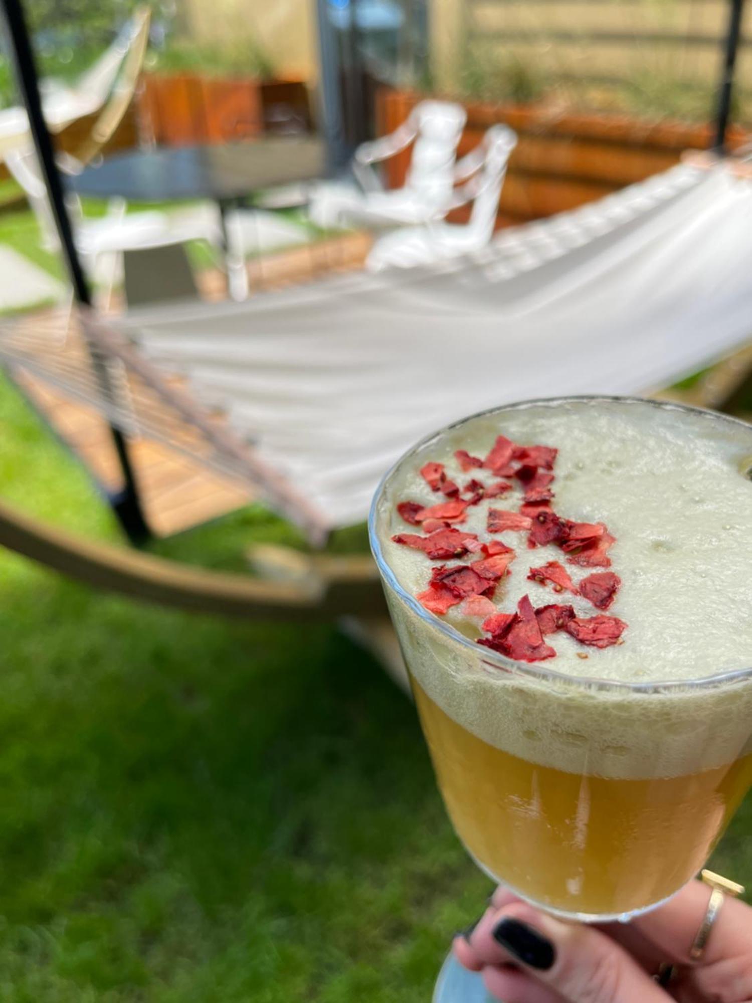 Royal Tulip Warsaw Centre Varsóvia Exterior foto The photo shows a close-up of a drink in a glass, topped with small pieces of red petals or dried flowers. The drink appears to have a frothy white layer above a golden-yellow liquid. In the background, there is a hammock set up on the grass, indicat