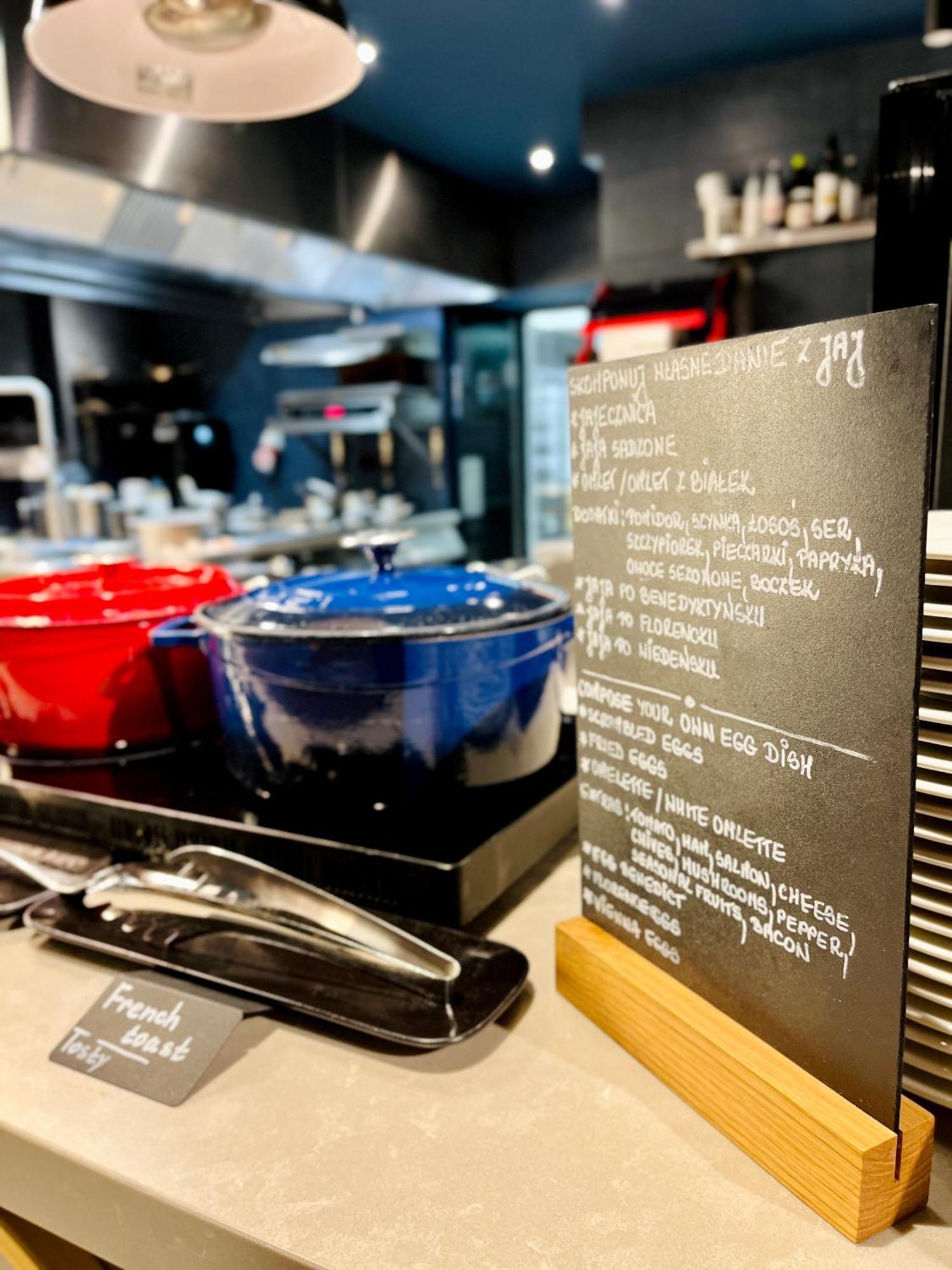 Royal Tulip Warsaw Centre Varsóvia Exterior foto The photo shows a modern kitchen setting with two large pots in the foreground—one red and one blue—placed on a countertop. Next to the pots, there is a menu or chalkboard listing various dishes. The board is decorated with handwritten notes in a lig