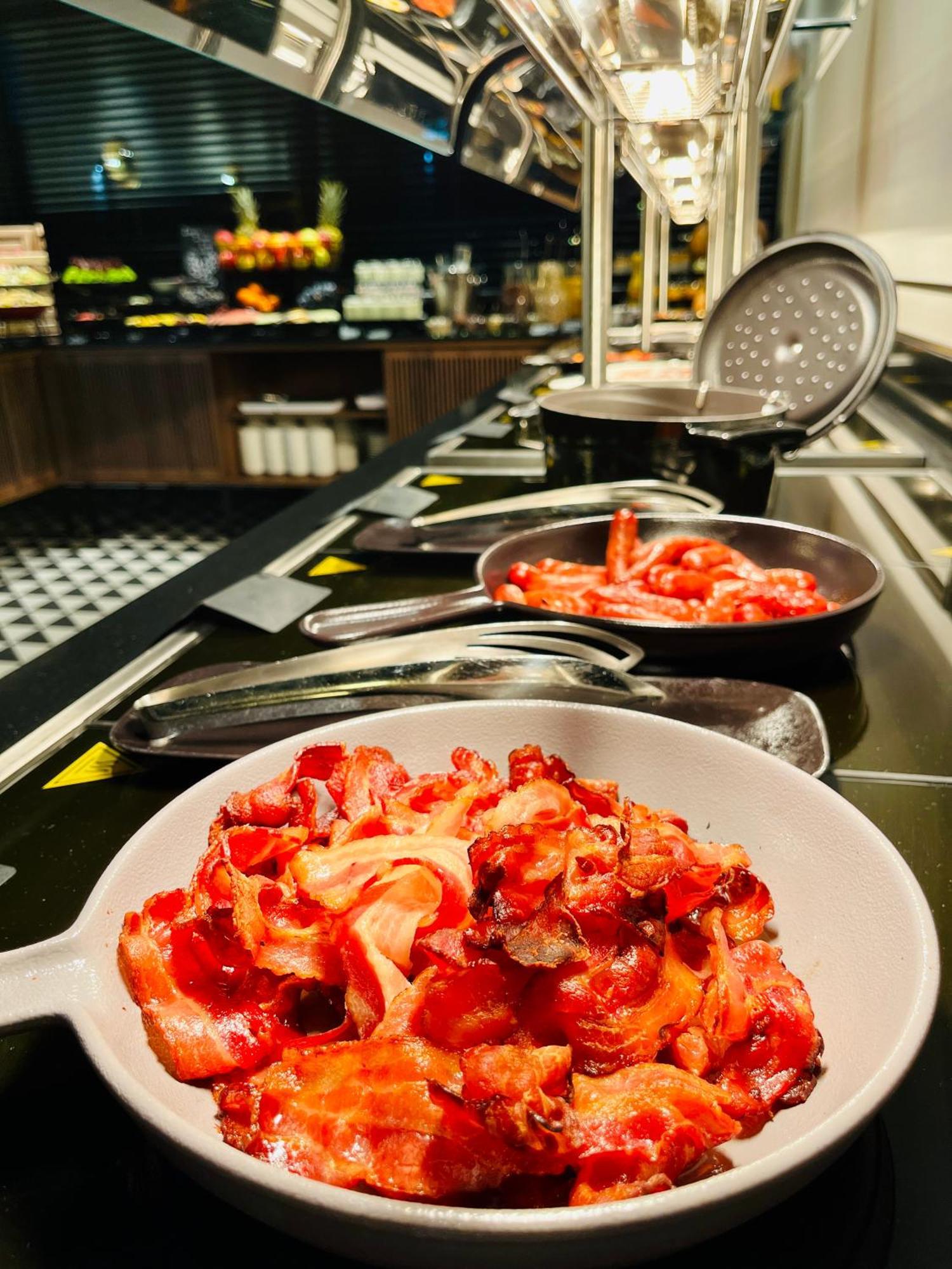 Royal Tulip Warsaw Centre Varsóvia Exterior foto The photo shows a selection of cooked bacon displayed on a buffet or serving station. There are several metal pans with more bacon strips, and one white bowl filled with an ample amount of bacon pieces, all appearing crispy and well-prepared. The set