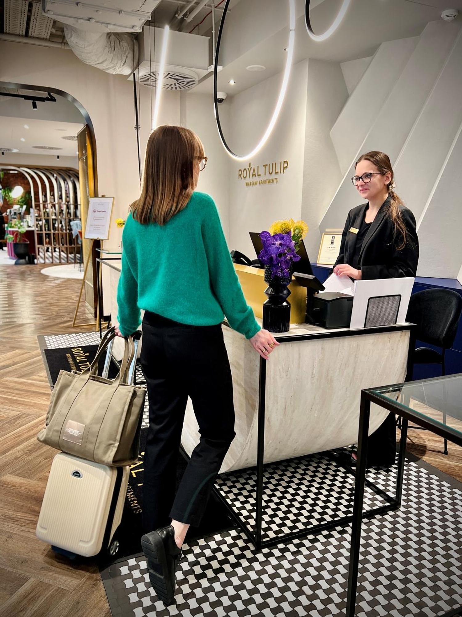 Royal Tulip Warsaw Centre Varsóvia Exterior foto The image shows a hotel reception area. A woman wearing a green sweater and black pants is standing at the reception desk, engaging with a staff member. The staff member, who is smiling and wearing a name tag, is standing behind the desk. There's a b