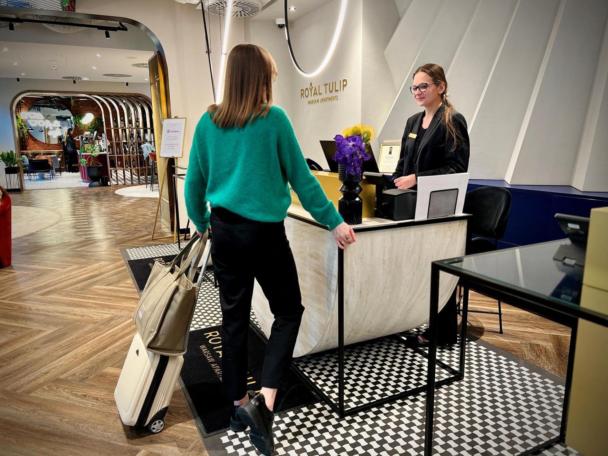Royal Tulip Warsaw Centre Varsóvia Exterior foto The photo depicts a hotel reception area. There is a young woman in a green sweater and black pants approaching the reception desk, carrying a suitcase and a bag. Behind the desk, another woman, likely a hotel staff member, is standing and appears to