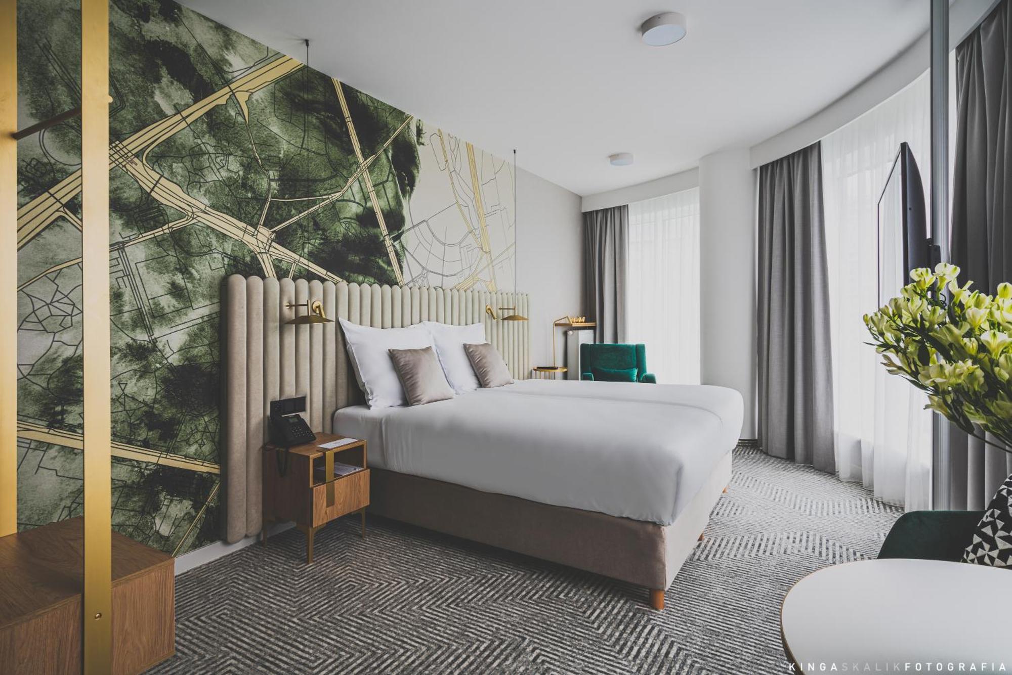 Royal Tulip Warsaw Centre Varsóvia Quarto foto The photo shows a modern hotel room featuring two beds with white bedding. The headboard is upholstered and elegantly designed. Behind the beds, there's a striking wall design with abstract green and gold patterns. The room has large windows with lig